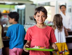 Portrait of smiling schoolboy holding food tray in canteen against classmates